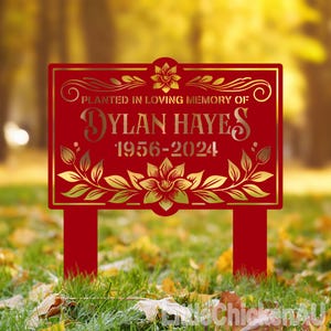 May include: Red metal garden stake with a gold inscription that reads "Planted in loving memory of Dylan Hayes 1956-2024". The inscription is surrounded by a gold floral design.