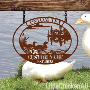 May include: A brown metal oval sign with a lake scene, ducks, and trees, hanging from chains. The sign has the text "CUSTOM TEXT", "CUSTOM NAME", and "EST.2023". Two white ducks are in the foreground.