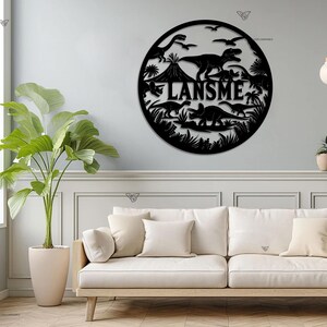 May include: Black metal wall art featuring a circle with dinosaurs and the name "LANSM E" in the center. The art is hanging on a white wall above a white couch.