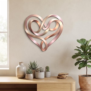 Può includere: Una scultura da parete a forma di cuore realizzata con un nastro continuo e fluido di metallo color oro rosa. Il design presenta intricati anelli sovrapposti che creano un senso di profondità e movimento, formando un cuore simmetrico.