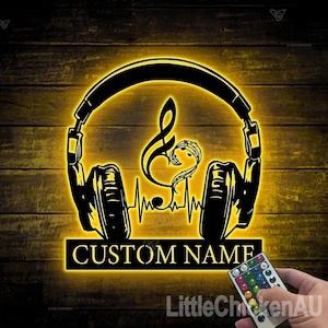 Könnte beinhalten: Ein schwarz-gelbes LED-Schild mit Kopfhörern, einer Musiknote und einer Herzschlaglinie. Das Schild trägt die Aufschrift "CUSTOM NAME" und leuchtet warm gelb. Eine Fernbedienung ist unten rechts zu sehen.