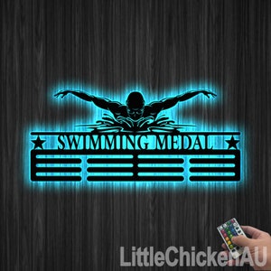 May include: A black metal medal display with a silhouette of a swimmer in a butterfly stroke. The display has the words "SWIMMING MEDAL" and five slots for medals. The display is illuminated with blue LED lights and comes with a remote control.