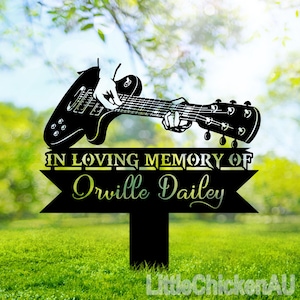 Könnte beinhalten: Schwarze Metallsilhouette einer Gitarre mit einer Hand, die darauf spielt. Die Gitarre ist auf einem schwarzen Metallpfahl montiert, an dem ein Schild mit der Aufschrift "In Loving Memory of Orville Dailey" angebracht ist.