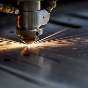 May include: Close-up of a laser cutter in operation, with bright orange sparks emanating from the cutting head. The machine is cutting a pattern into a metal sheet. The focus is on the precision and heat of the process.