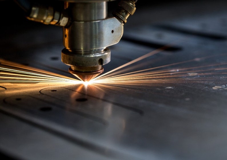 May include: Close-up of a laser cutter in operation, with bright orange sparks emanating from the cutting head. The machine is cutting a pattern into a metal sheet. The focus is on the precision and heat of the process.