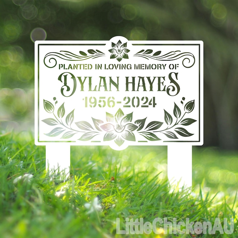 May include: White metal garden sign with a floral design and the text "Planted in loving memory of Dylan Hayes 1956-2024".