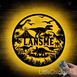 May include: A black metal circle with a dinosaur theme. The circle has a silhouette of a volcano, a T-Rex, a Triceratops, and other dinosaurs. The name "LANSMEN" is written in the center of the circle. The circle is illuminated with a yellow light.