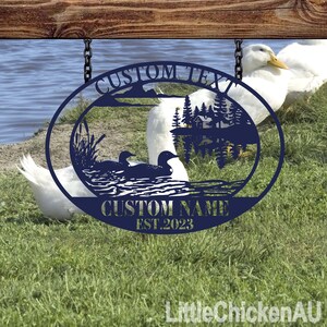 May include: A navy blue metal sign with a lake scene featuring two loons, trees, and a cabin. The sign has the text "CUSTOM TEXT" at the top and "CUSTOM NAME EST.2023" at the bottom. The sign is hanging from a wooden beam with chains.