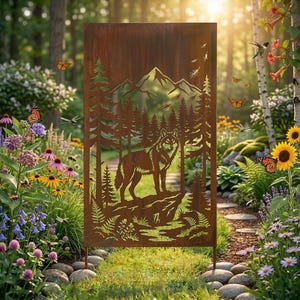 Puede incluir: Pantalla de jardín rectangular de metal marrón oxidado con un diseño recortado de un lobo en un bosque con montañas al fondo. La pantalla está sostenida por dos estacas de metal y está rodeada por un exuberante jardín con flores y mariposas.