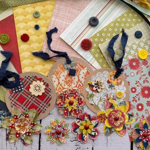May include: A collection of circular paper tags and layered paper flowers in various patterns and colors, including red, yellow, green, and blue. The tags feature dark blue ribbon ties, and several small plastic buttons are scattered across the surface.