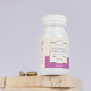 Op de afbeelding: Een witte fles voedingssupplementcapsules met een gouden etiket en de tekst "RAUDONŲJŲ DOBILŲ ŽIEDAI RED CLOVER BLOSSOMS". Twee capsules liggen naast de fles. De fles bevat 90 capsules.