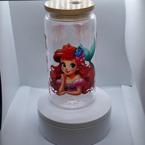 May include: A clear glass tumbler with a bamboo lid featuring a colorful illustration of a red-haired mermaid with a crown and a blue fish. The tumbler has a pink and purple design.
