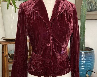 May include: A burgundy velvet jacket with a classic collar and button closure. The jacket features gathered detailing on the shoulders and pockets, and is displayed on a mannequin. The jacket is a rich, deep red color.