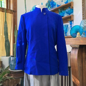 May include: A vibrant blue blazer with a mandarin collar and a single button closure. The jacket is displayed on a mannequin, paired with a gray skirt. The background features decorative glass items and plants.