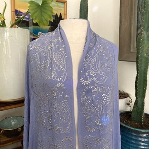 May include: A periwinkle blue scarf with intricate silver embellishments in a paisley pattern. The scarf is draped over a mannequin, showcasing its design. The fabric appears lightweight and flowing, suggesting a delicate texture.