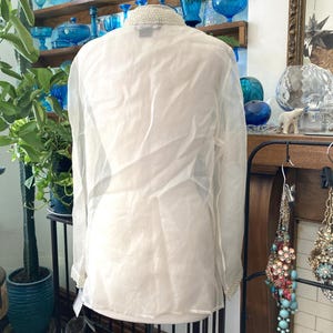May include: A sheer white blouse with long sleeves and a beaded collar and cuffs. The blouse is displayed on a mannequin. The background includes a shelf with blue glass objects and decorative items.
