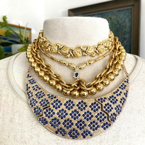 May include: A collection of gold-toned necklaces displayed on a mannequin. The necklaces include a floral design, a chain with a small pendant, and a statement necklace with blue gemstone accents. The necklaces are layered for a bold look.