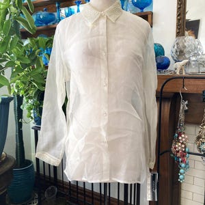 May include: A sheer white button-down shirt with long sleeves. The collar and cuffs are embellished with small, white beads. The shirt is displayed on a mannequin, showcasing its delicate fabric and design.