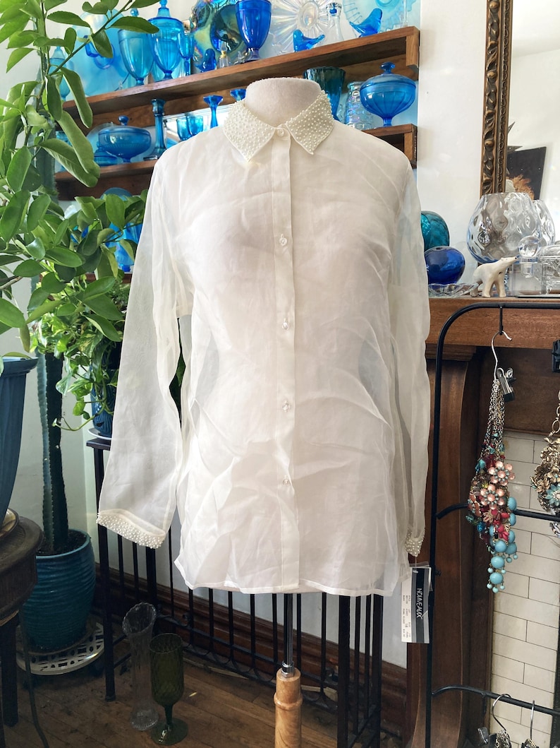 May include: A sheer white button-down shirt with long sleeves. The collar and cuffs are embellished with small, white beads. The shirt is displayed on a mannequin, showcasing its delicate fabric and design.
