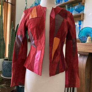 May include: A red suede jacket with a patchwork design. The jacket features a collarless design and long sleeves. The patchwork includes shades of red, orange, and brown. The jacket is displayed on a mannequin.