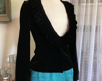 May include: A black velvet jacket with a ruffled collar is displayed on a mannequin. The jacket has long sleeves and a tailored fit. The mannequin is paired with a turquoise skirt. The jacket is a classic piece of apparel.