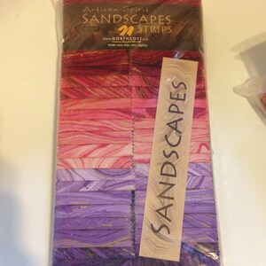 May include: A clear plastic package containing a stack of fabric strips labeled Artisan Spirit SANDSCAPES STRIPS by Northcott. The strips feature a gradient of colors from deep red and pink to light pink, lavender, and dark purple with metallic accents.