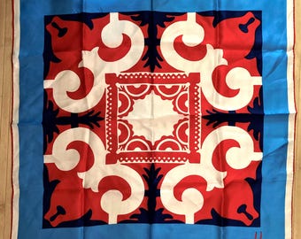 May include: Square silk scarf with a vibrant geometric design. The scarf features a central white square with red and white patterns, surrounded by red and navy shapes. A light blue border frames the design, with the name "Usna" in the corner.