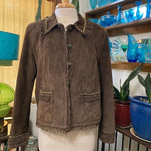 May include: A dark brown suede jacket featuring a pointed collar with metallic studs, a front button closure, and two front pockets with stud detailing. The jacket is finished with fringe trim along the collar, front opening, hem, and cuffs.