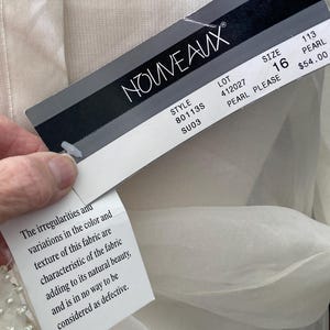 May include: A close-up of a tag from the brand NOUVEAUX, attached to a cream-colored garment. The tag displays the brand name, style, size 16, and other details. A smaller tag with text about the fabric's texture is also visible.