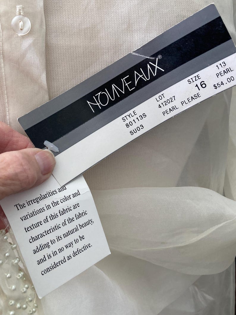 May include: A close-up of a tag from the brand NOUVEAUX, attached to a cream-colored garment. The tag displays the brand name, style, size 16, and other details. A smaller tag with text about the fabric's texture is also visible.