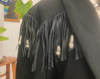 May include: Black jacket with leather fringe detailing across the shoulders. The fringe is accented with silver-toned metal beads. The jacket has a classic collar and a tailored fit, suggesting a vintage or bohemian style.