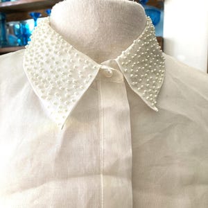 May include: Close-up of a white blouse with a Peter Pan collar. The collar is embellished with small, white, pearl-like beads. The blouse has a button-down front and is made of a sheer, lightweight fabric.