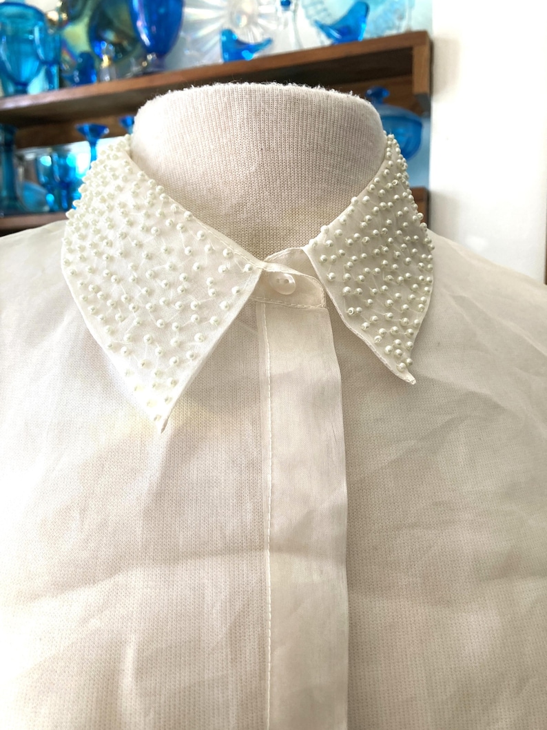 May include: Close-up of a white blouse with a Peter Pan collar. The collar is embellished with small, white, pearl-like beads. The blouse has a button-down front and is made of a sheer, lightweight fabric.