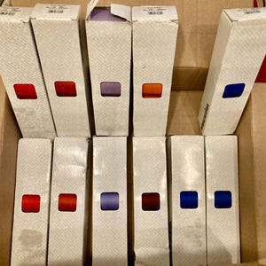 Puede incluir: Once cajas blancas altas y rectangulares dispuestas en dos filas, cada una con un pequeño recorte cuadrado que revela una cinta de diferente color en su interior. Los colores visibles a través de los recortes incluyen tonos de rojo, naranja, morado y azul.