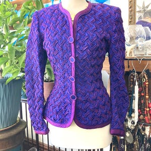 May include: A vintage purple jacket with a textured woven design. The jacket features a round collar, three buttons, and purple trim. The sleeves are long, and the jacket is displayed on a mannequin.