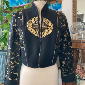 May include: Black jacket with gold embroidered details. The jacket features a high collar and a central gold emblem. Gold bird patterns adorn the sleeves. The jacket is displayed on a white mannequin.