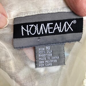 May include: A close-up of a tag from a garment. The tag is black with white text that reads "NOUVEAUX". Below the brand name, a smaller tag indicates the size is 16, made of 100% silk, and made in China. The garment is a light cream color.