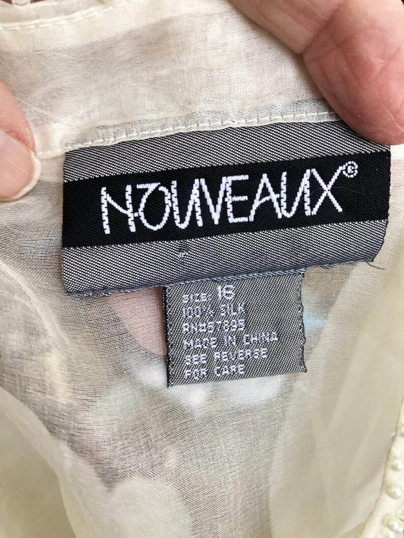May include: A close-up of a tag from a garment. The tag is black with white text that reads "NOUVEAUX". Below the brand name, a smaller tag indicates the size is 16, made of 100% silk, and made in China. The garment is a light cream color.