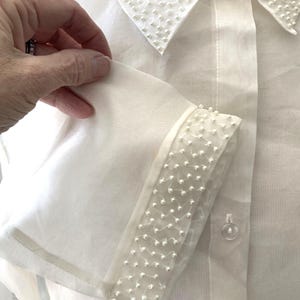 May include: A white button-down shirt with a sheer, long sleeve. The collar and sleeve cuffs are embellished with small, white beads. The shirt has a classic, elegant design.