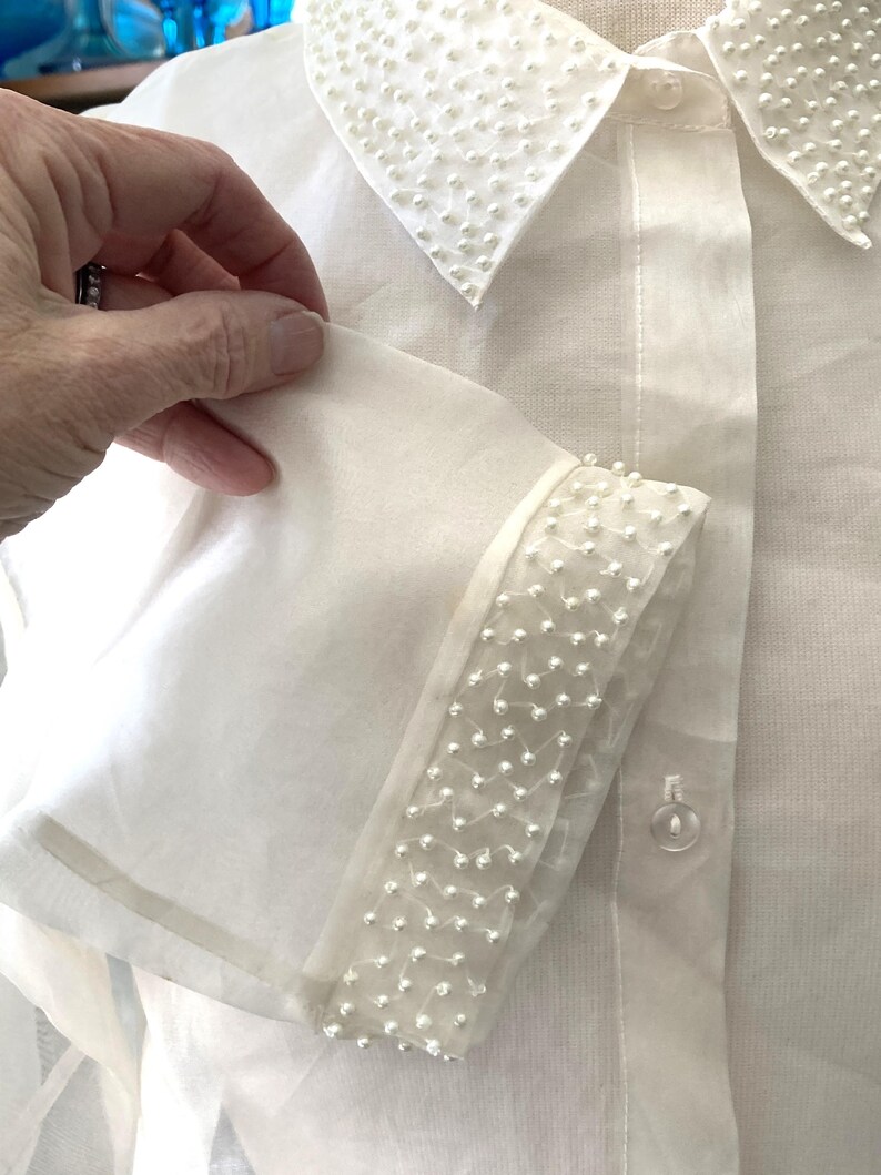 May include: A white button-down shirt with a sheer, long sleeve. The collar and sleeve cuffs are embellished with small, white beads. The shirt has a classic, elegant design.