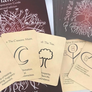 May include: A set of tarot cards with a brown book titled "Nanrunes Interpretation Guide, Second Edition" by Molly Remer and Shekhinah Mountainwater. The cards feature illustrations of a crescent moon, a tree, and a woman with a tree growing from her head. The cards are laid out in a spread with the text "Flying Woman Layout" visible.