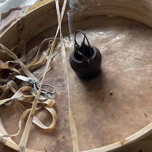 Peut inclure: Un tambour à cadre en bois avec une peau de tambour en cuir blanc. Un petit brûleur d'encens en métal est posé sur la peau du tambour, avec de la fumée qui s'en échappe. Une paire de ciseaux et un élastique sont également sur la peau du tambour.