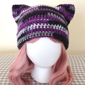 May include: A handmade crochet cat ear hat in shades of purple, black, gray, and white. The hat is designed with a striped pattern and features two pointed ears at the top. The hat is displayed on a white mannequin head with pink hair.