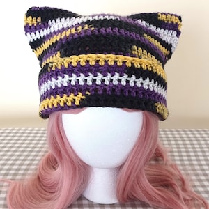 May include: A handmade crochet cat ear hat in black, gold, white, and purple. The hat is displayed on a white foam head with pink hair, set against a checkered background. The hat's design features alternating stripes of color.