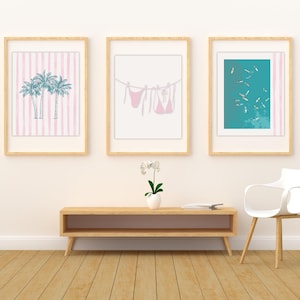 May include: Three framed art prints with a beach theme. One print features palm trees on a pink striped background. Another shows a pink bikini hanging on a clothesline. The third depicts swimmers in a teal pool.