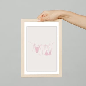 May include: A framed print featuring a minimalist line drawing of three pink bikinis hanging on a clothesline. The artwork is set against a light beige background, with a simple, clean aesthetic. The frame is made of light-colored wood.