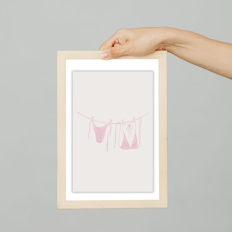 May include: A framed print featuring a minimalist line drawing of three pink bikinis hanging on a clothesline. The artwork is set against a light beige background, with a simple, clean aesthetic. The frame is made of light-colored wood.