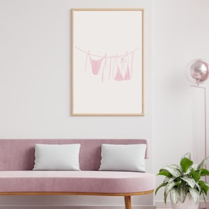 May include: A framed print featuring a minimalist line drawing of a pink bikini set hanging on a clothesline. The artwork is set against a white background, framed in a light wood. The print is displayed above a pink sofa with white pillows.