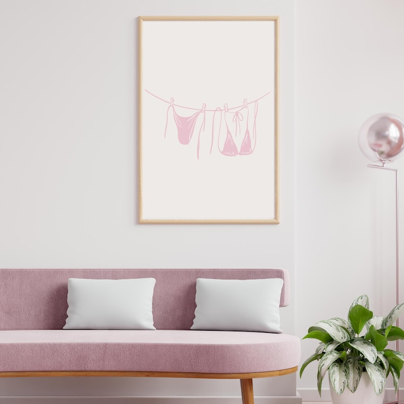 May include: A framed print featuring a minimalist line drawing of a pink bikini set hanging on a clothesline. The artwork is set against a white background, framed in a light wood. The print is displayed above a pink sofa with white pillows.