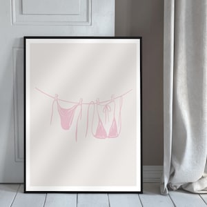 May include: A framed print featuring a pink line drawing of swimwear hanging on a clothesline. The minimalist art piece includes a bikini top, bottoms, and a one-piece swimsuit, all in a soft pink hue. The artwork is set against a neutral background.